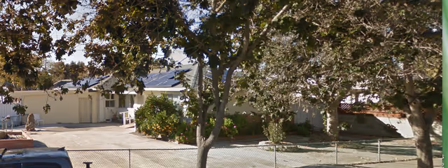 Exterior view of a single-story building partially obscured by trees with green leaves. The building has solar panels on the roof and is surrounded by a paved area and some bushes. A chain-link fence is visible in the foreground.