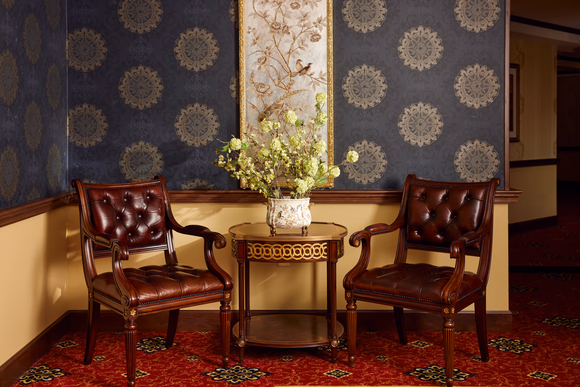 Two dark brown leather armchairs with button tufting are positioned on either side of a small round wooden table with decorative gold accents. A vase with green and white flowers sits on the table. The background features a dark blue wallpaper with gold circular patterns and a framed artwork with floral and bird motifs. The floor is covered with a red carpet with intricate patterns.