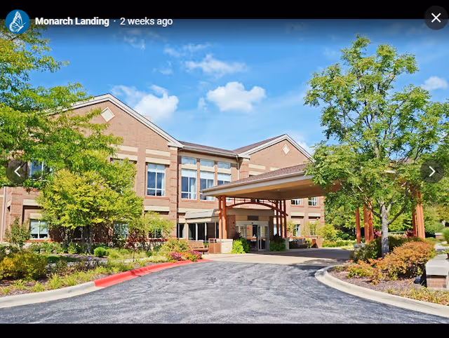 Exterior view of Monarch Landing senior living facility showing a two-story brick building with large windows, a covered entrance with wooden beams, surrounded by landscaped greenery and trees under a blue sky with some clouds.