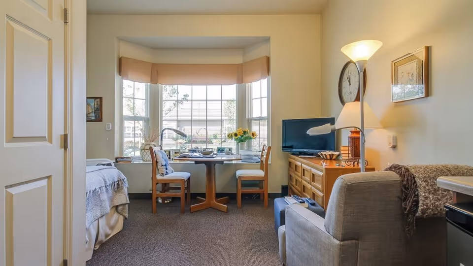 A cozy assisted living room with a bed on the left, a round wooden table with two chairs in front of a large bay window, a wooden dresser with a TV and lamp on the right, and a comfortable armchair with a blanket draped over it. The room is well-lit with natural light coming through the window.