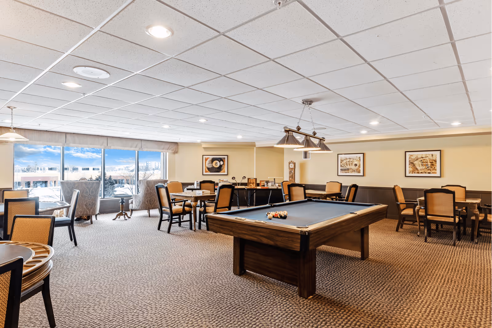 A spacious recreational room with a pool table in the center, surrounded by multiple tables and chairs. Large windows on one side let in natural light and offer a view of buildings and trees outside. The room has a carpeted floor, beige walls with framed pictures, and a ceiling with recessed lighting. There are also a couple of armchairs near the windows.