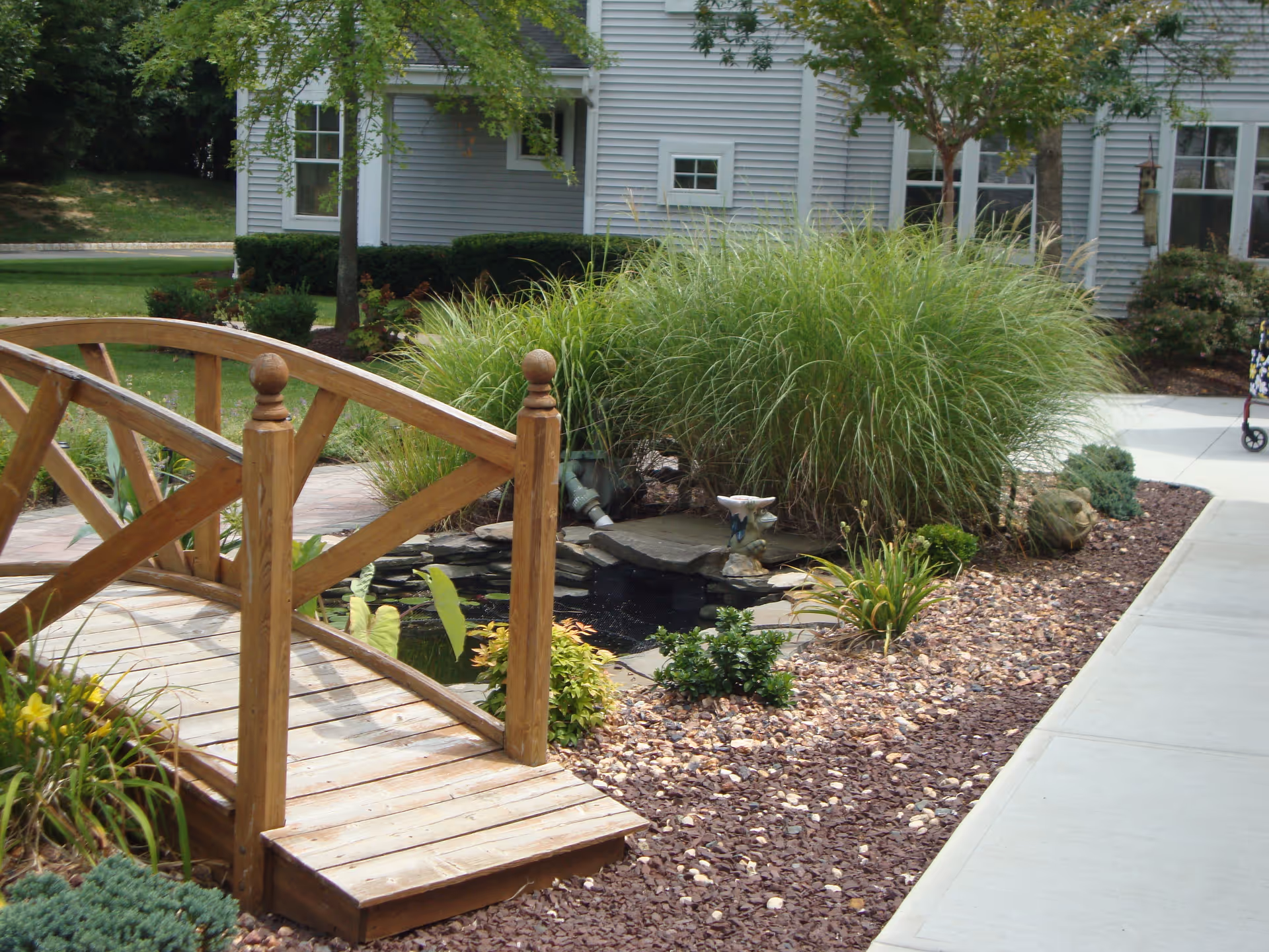A small wooden footbridge over a garden pond surrounded by various plants and shrubs, with a gray building in the background and a concrete walkway on the right side.