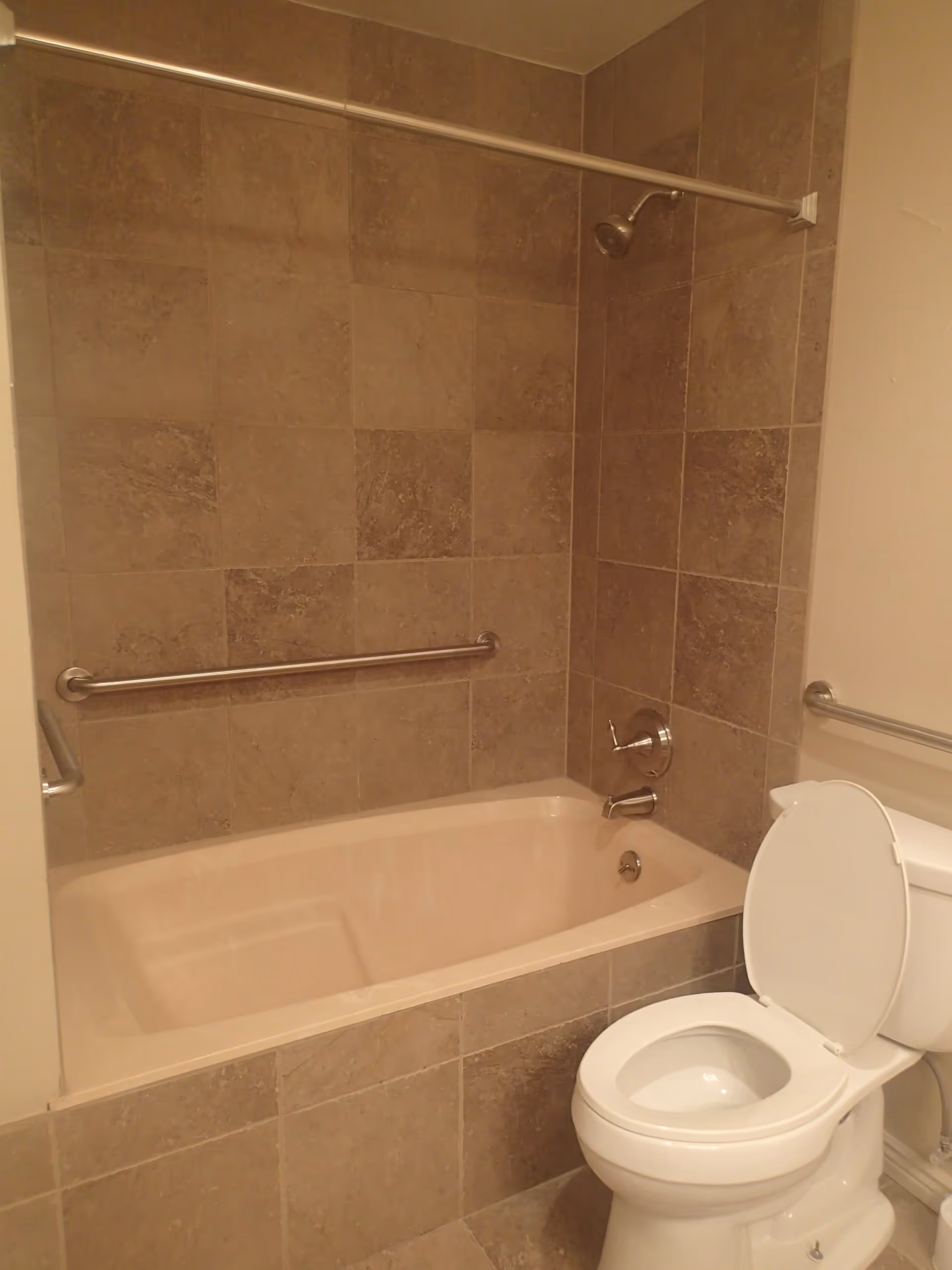 A bathroom featuring a bathtub with beige tiles on the walls and a white toilet with the lid open. There are metal grab bars installed on the wall next to the bathtub and beside the toilet for support.