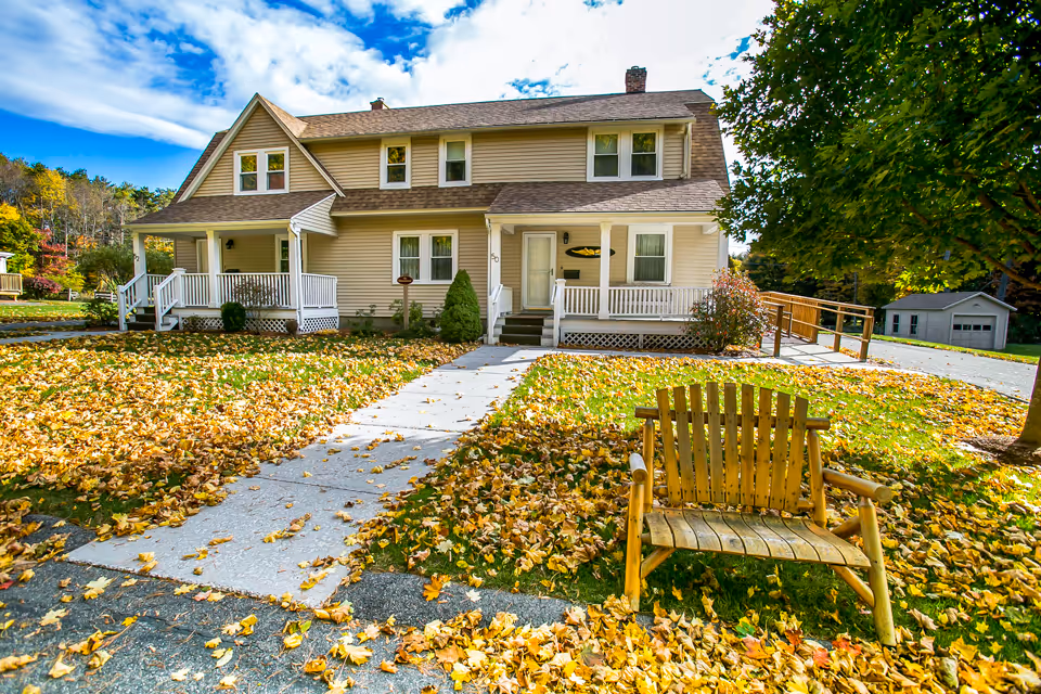 Two-story beige assisted living house with front porch, walkway, and autumn leaves.