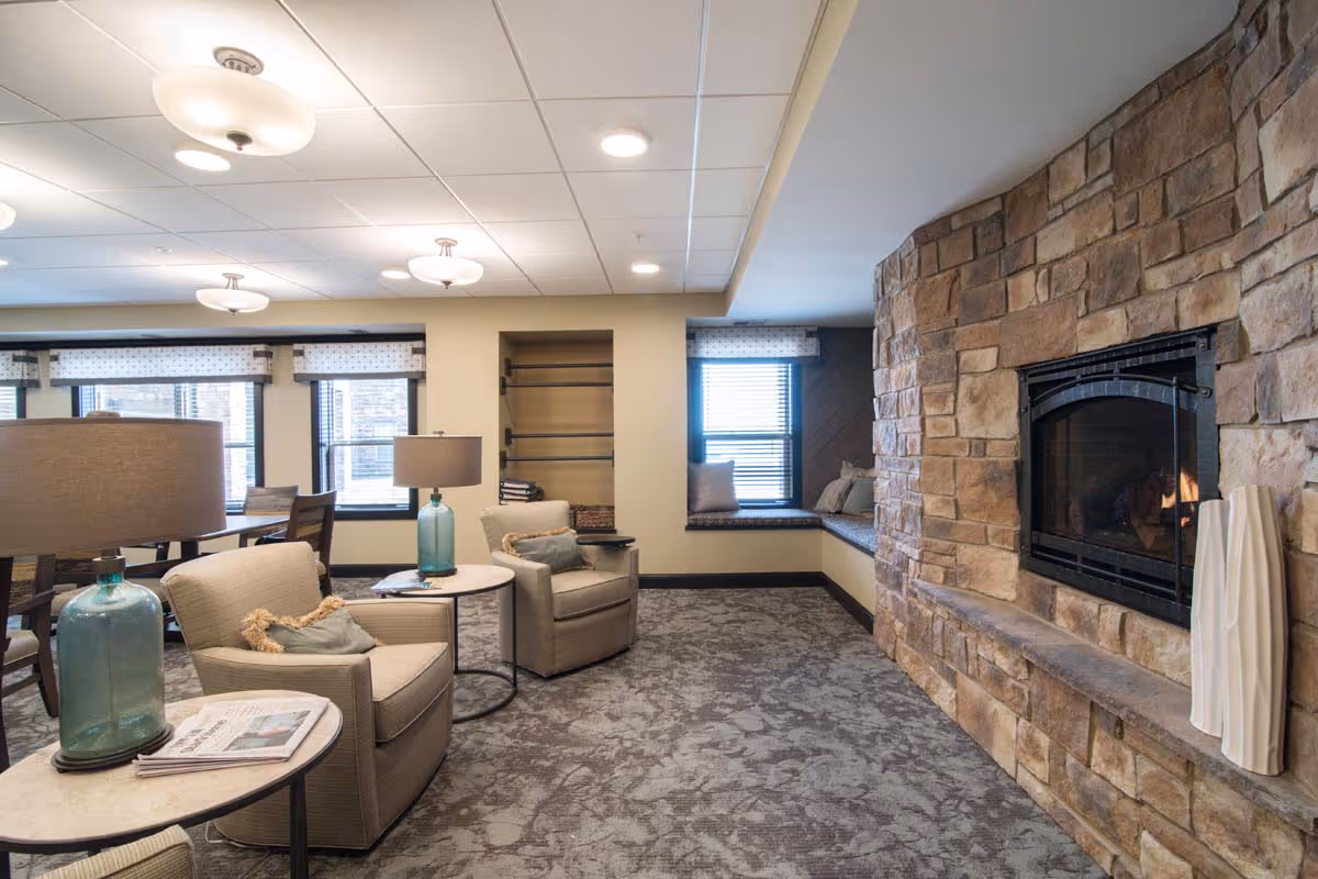 Cozy communal lounge with armchairs, side tables and lamps facing a stone fireplace and window seat.