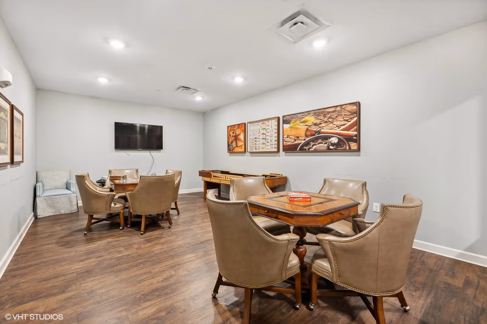 Communal game room with card tables, leather chairs, a wall-mounted TV and a shuffleboard table.