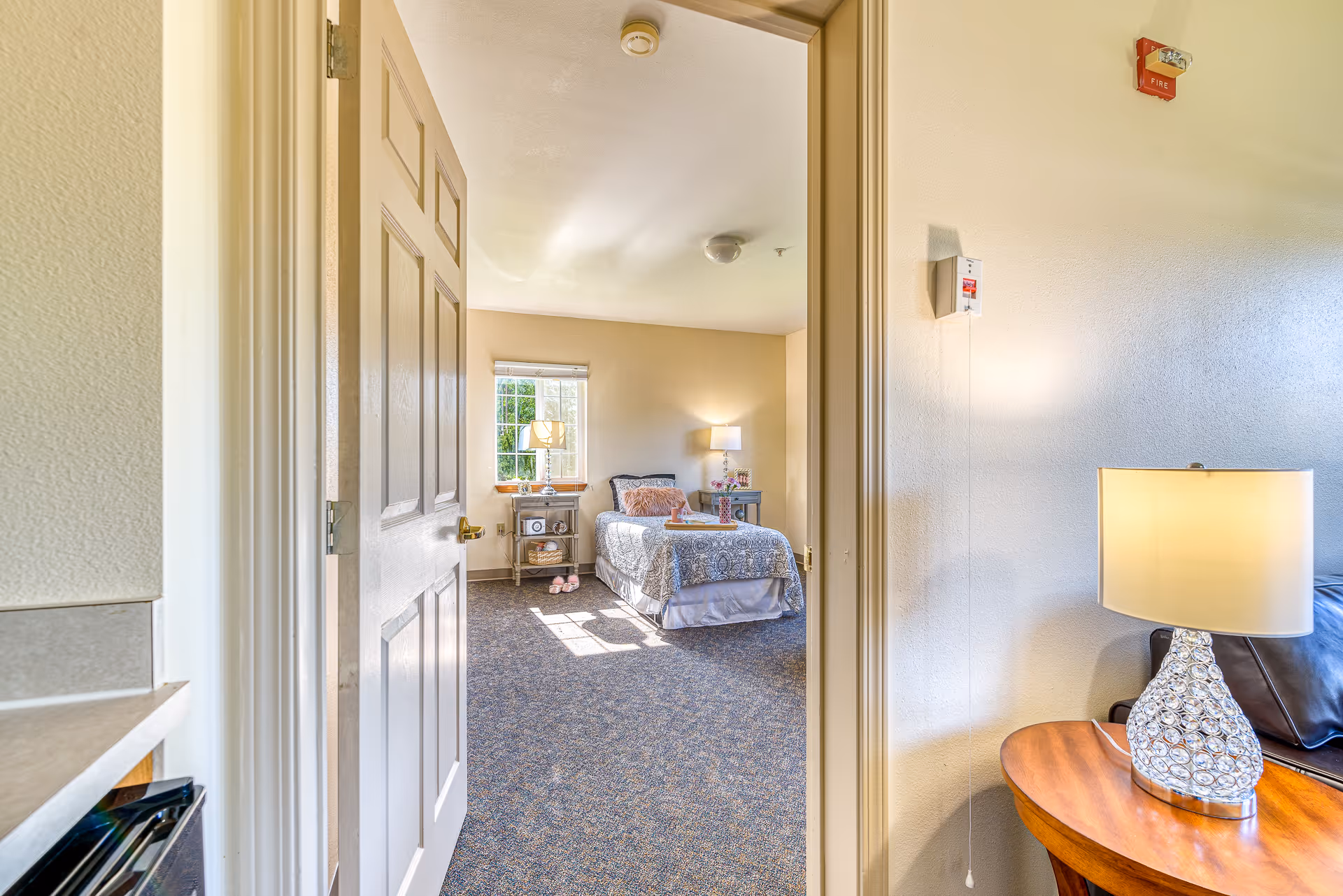 View through an open door into a bedroom with a single bed covered in a patterned bedspread, a bedside table with a lamp, and a window letting in natural light. In the foreground, part of a living area is visible with a wooden side table and a decorative lamp.