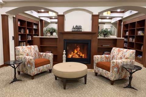 A cozy living room area with two patterned armchairs featuring orange cushions, positioned on either side of a round beige ottoman. Behind the ottoman is a fireplace with a warm fire burning, flanked by built-in wooden bookshelves filled with books and decorative plants. The room has a neutral carpet and soft lighting.