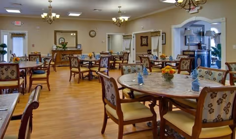 A spacious dining room with multiple round wooden tables and cushioned chairs arranged neatly. Each table has a small floral centerpiece and folded blue napkins. The room features hardwood flooring, beige walls, chandeliers, and a large mirror above a wooden sideboard. There are windows and doors along the walls, allowing natural light to enter.