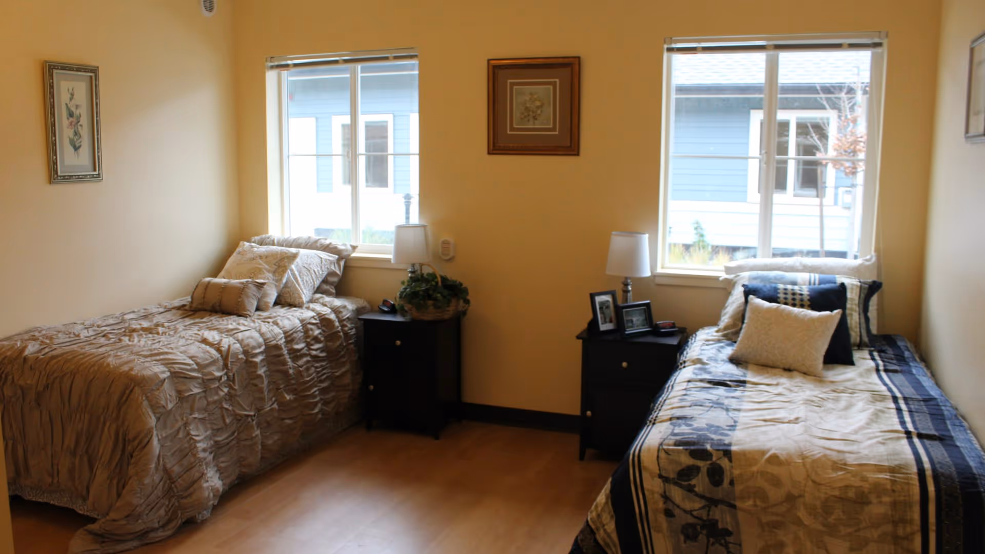 A cozy bedroom with two single beds placed against opposite walls. Each bed has decorative pillows and a bedside table with a lamp. Two windows allow natural light into the room, and framed artwork hangs on the walls.