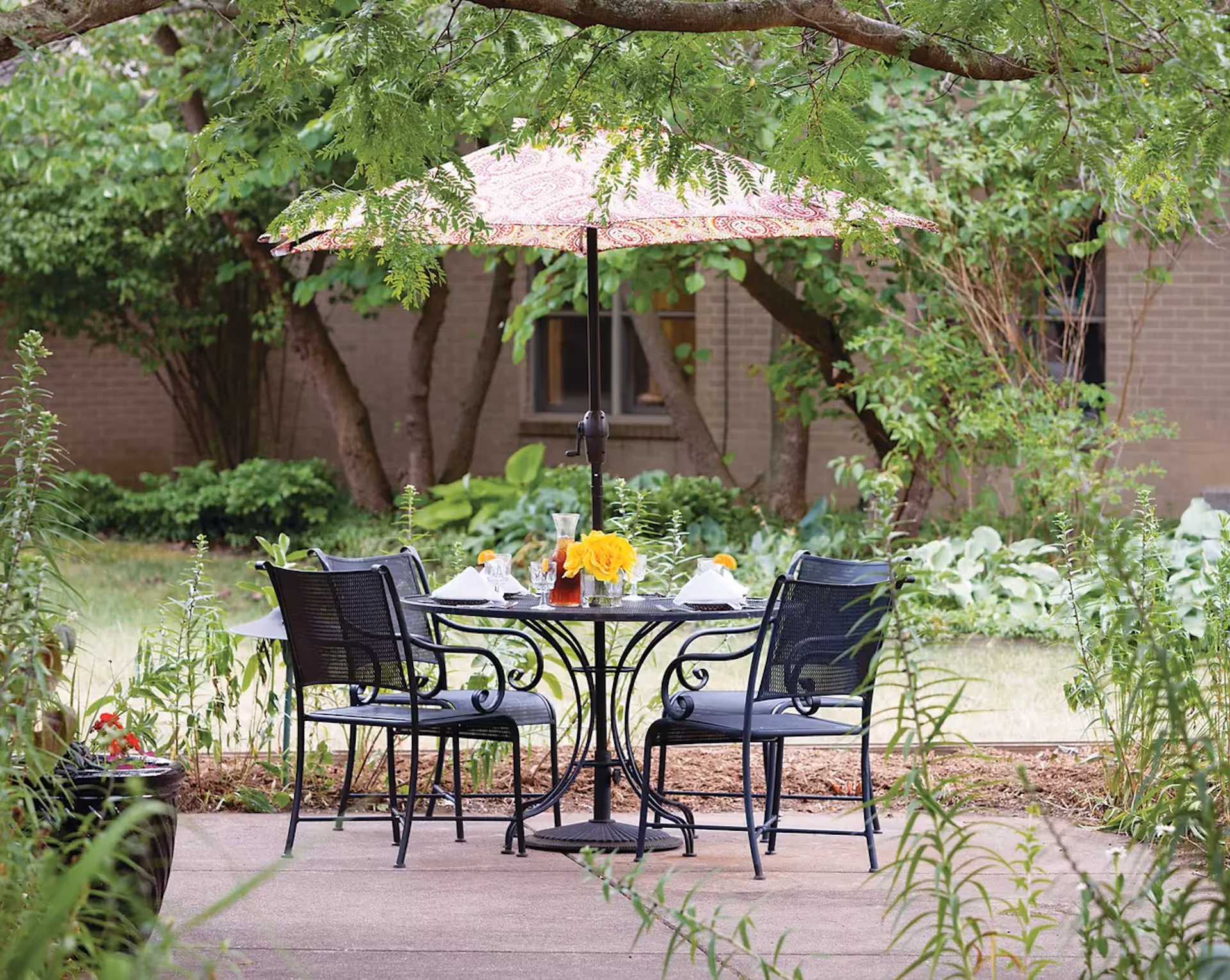 Outdoor patio area with a round black metal table and four matching chairs. The table is set with a pitcher of iced tea, glasses, napkins, and a small vase of yellow flowers. A large patterned umbrella provides shade, and the area is surrounded by green plants and trees with a brick building in the background.