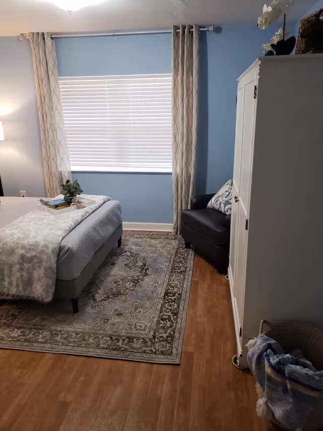 A cozy bedroom with light blue walls and a large window covered by white blinds and beige patterned curtains. The room features a bed with a gray upholstered frame, a patterned blanket, and a tray with books and a small plant on it. There is a dark armchair with a decorative pillow in the corner next to a white wardrobe. The floor is wooden with a large ornate area rug under the bed. A basket with folded blankets is placed near the wardrobe.