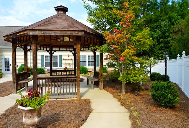A wooden gazebo surrounded by lush greenery and colorful plants at Lancaster Grove Senior Living, with a paved pathway leading to the entrance.