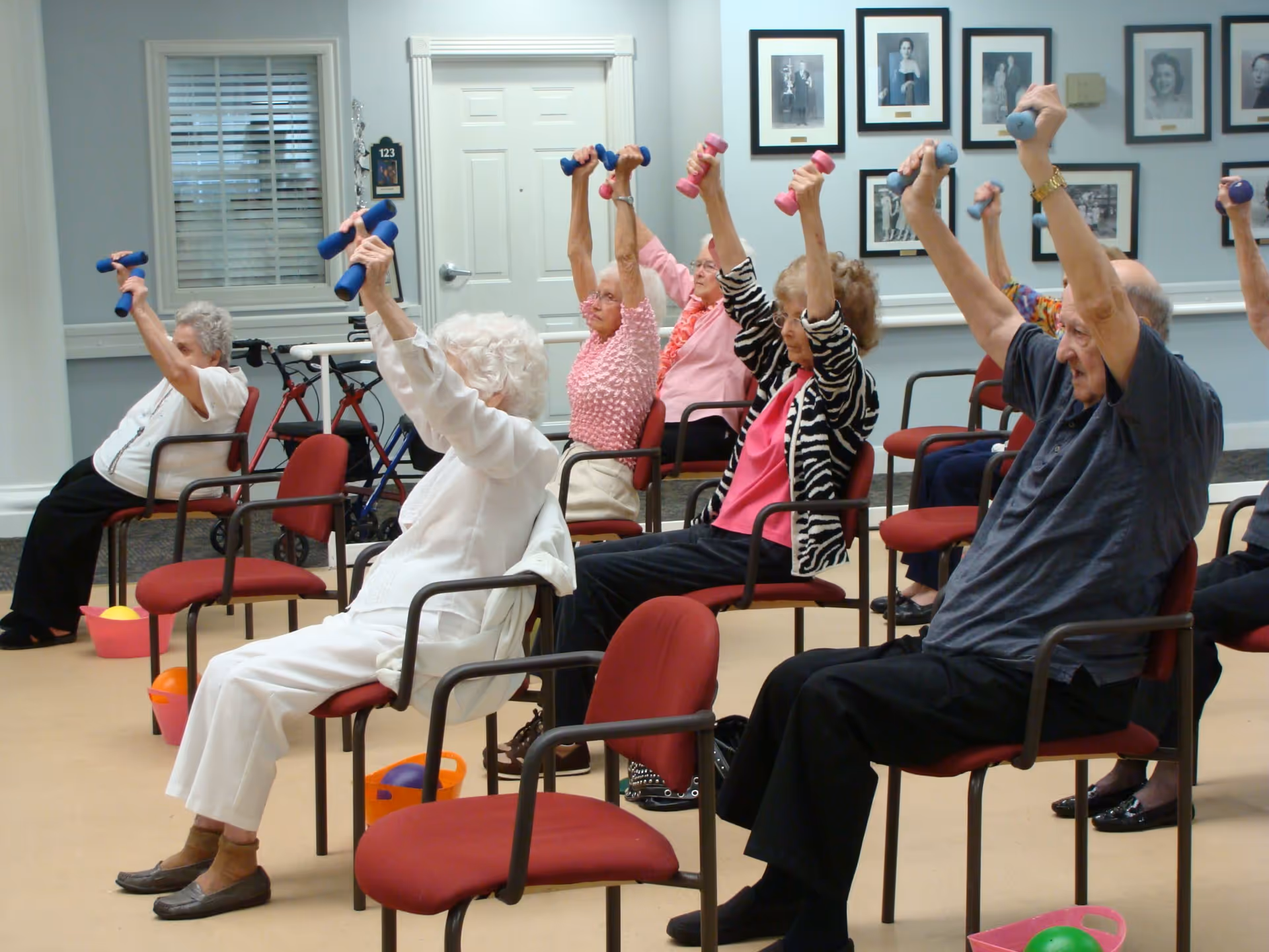 A group of elderly individuals seated in chairs in a room, participating in a seated exercise class by lifting small dumbbells above their heads. The room has light blue walls adorned with framed black and white portraits, and there are walkers and colorful exercise balls on the floor.