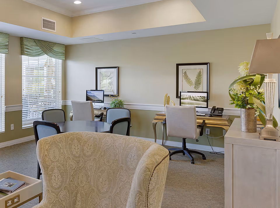 Bright communal seating area with round table, armchairs and two computer workstations against a wall with framed botanical prints.