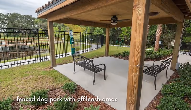 A covered outdoor seating area with two metal benches on a concrete slab, surrounded by a fenced dog park with grassy areas and trees in the background. There is a ceiling fan under the wooden roof structure and signage on the fence.