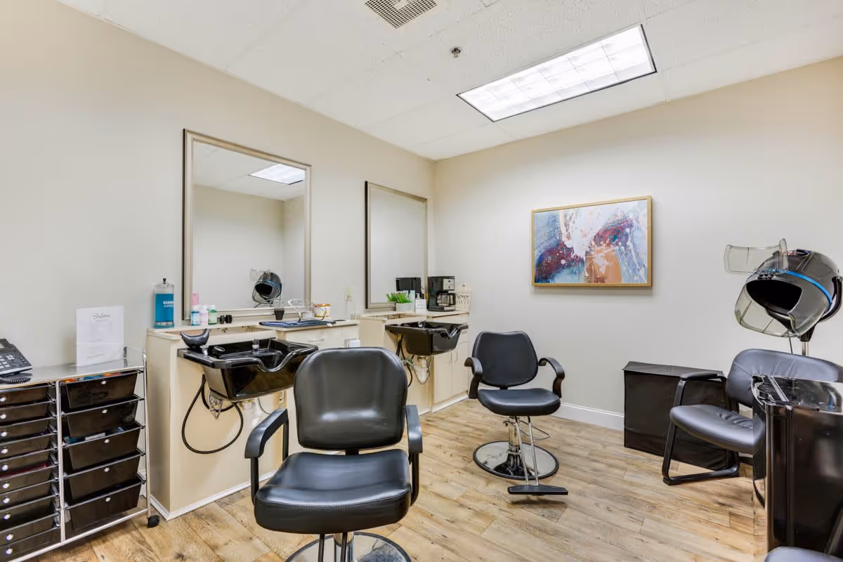 Interior view of a hair salon area with two black salon chairs in front of two sinks and mirrors. There is a hair dryer chair on the right side, a small black storage cabinet, and a colorful abstract painting on the wall. The floor is wooden, and the walls are light-colored.