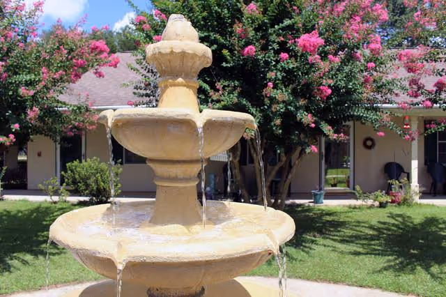A beige three-tiered water fountain with water flowing from each tier, set in a grassy courtyard with blooming pink flowering trees and a single-story building with a covered walkway in the background.