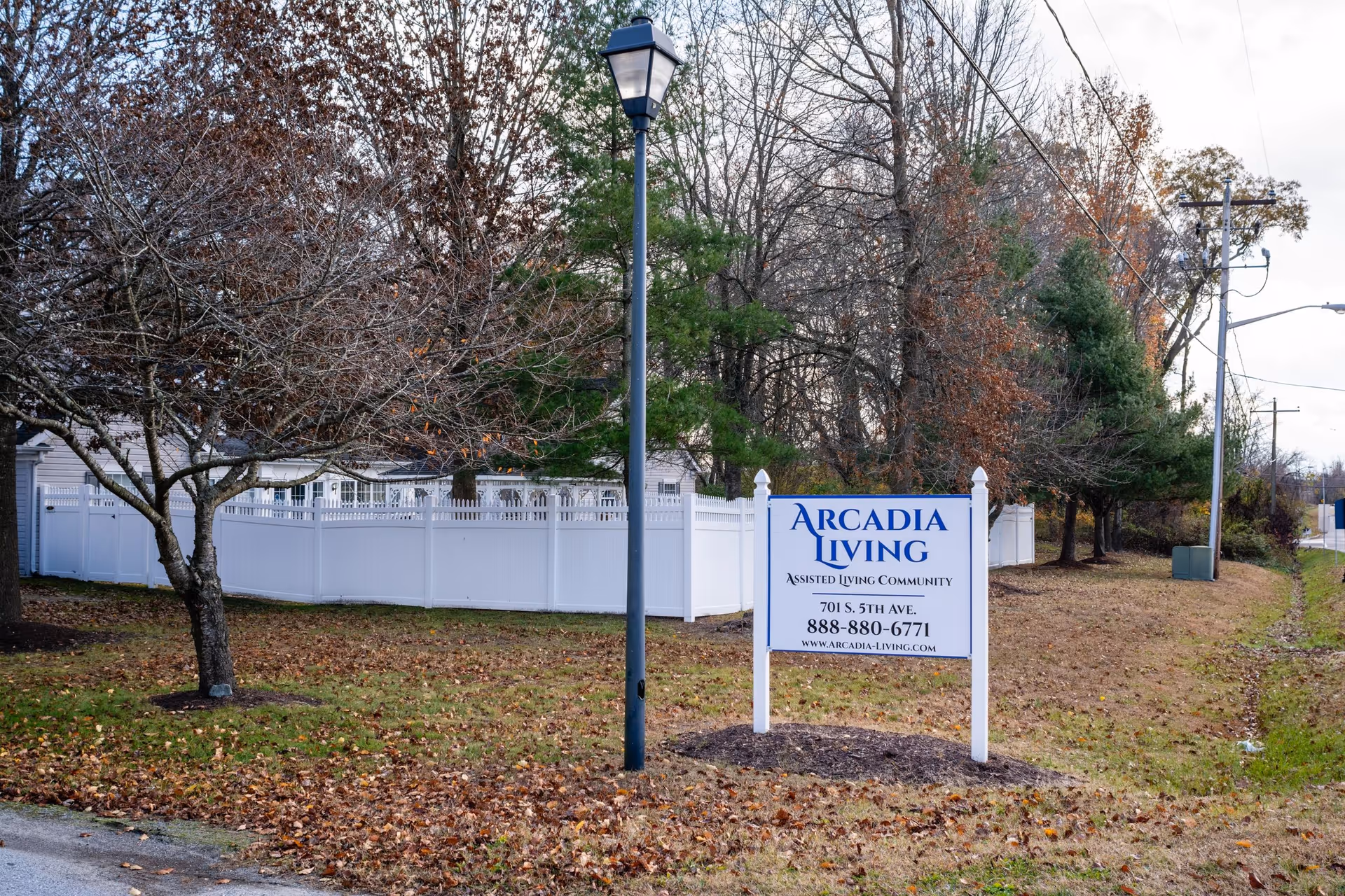 A roadside lawn with a white sign for Arcadia Living in front of a white fence and leafless trees.