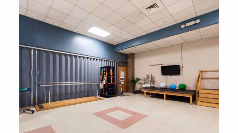 A rehabilitation room with parallel bars for walking exercises, a rack holding various colorful weights, a bench with exercise balls, a wall-mounted TV, and wooden stairs for physical therapy. The room has a tiled ceiling and a floor with pink square patterns.