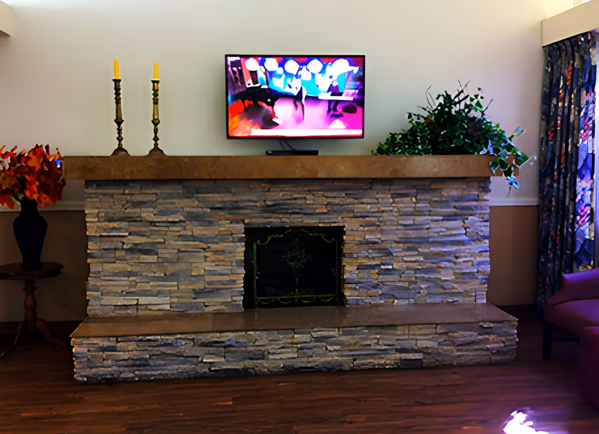 A stone fireplace with a wooden mantel in a living room. On the mantel, there are two tall candlesticks with yellow candles and a green leafy plant. Above the mantel, a flat-screen TV is mounted on the wall displaying a colorful image. To the left of the fireplace, there is a small wooden table with a black vase holding red flowers. To the right, part of a purple armchair and floral curtains are visible. The floor is wooden.