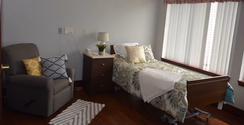 A small bedroom with a single hospital-style bed covered with a patterned bedspread and pillows. Next to the bed is a dark wooden nightstand with a lamp, a small plant, and a telephone. To the left of the nightstand is a gray recliner chair with two decorative pillows, one yellow and one gray with white patterns. The floor is wooden, and there is a small striped rug in front of the chair. A window with vertical blinds lets in natural light on the right side of the room.