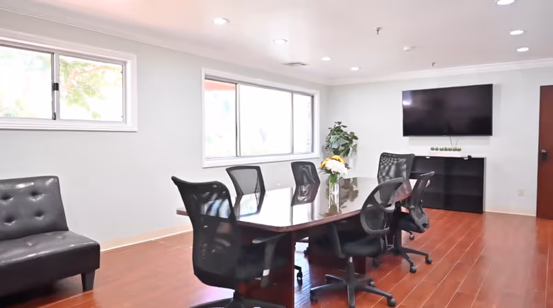 A bright conference room with a long wooden table surrounded by six black mesh office chairs. There is a black leather chair against the left wall, two large windows letting in natural light, a flat screen TV mounted on the wall, a black cabinet beneath the TV, and a potted plant in the corner. A vase with flowers is placed on the table.