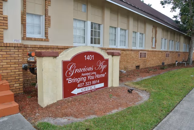 Exterior view of the Gracious Age assisted living facility showing a brick and beige building with multiple windows and a red sign in front that reads '1401 Gracious Age Assisted Living Bringing You Home' with a phone number and an arrow pointing to the office.