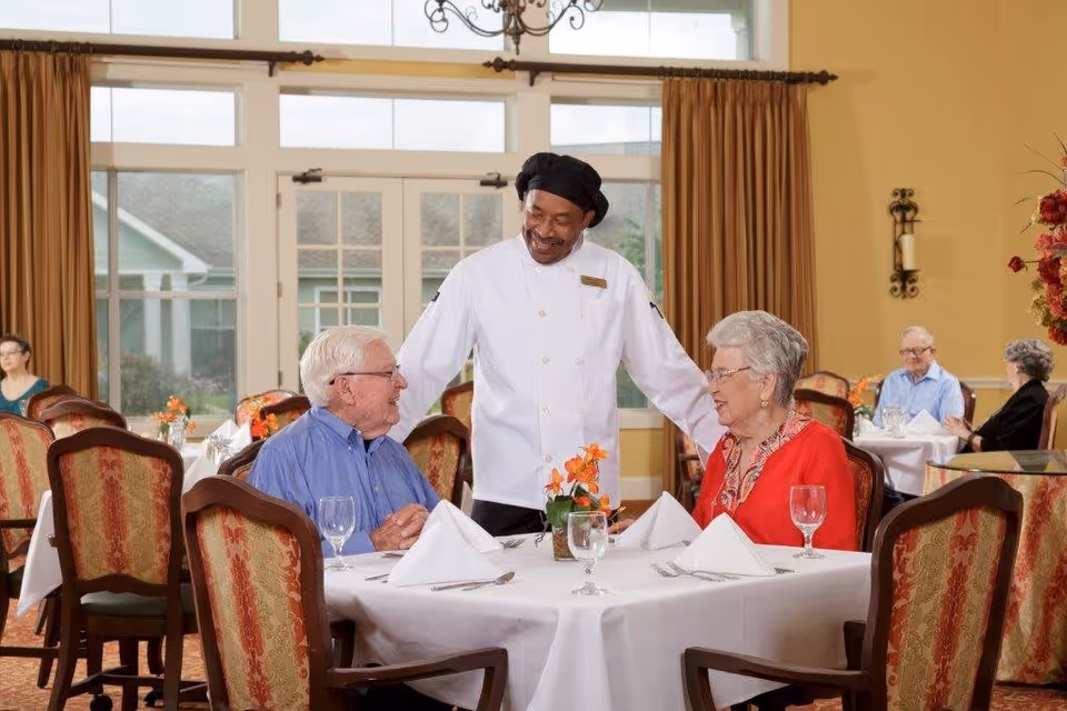 A chef in a white uniform and black hat stands smiling and talking to an elderly man and woman seated at a dining table set with white tablecloth, napkins, and glassware in a bright dining room with large windows and brown curtains.