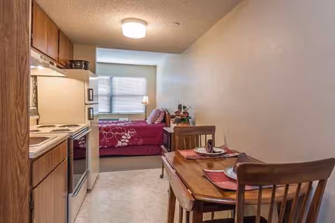 Compact studio senior living apartment with kitchenette, dining table, and bed.