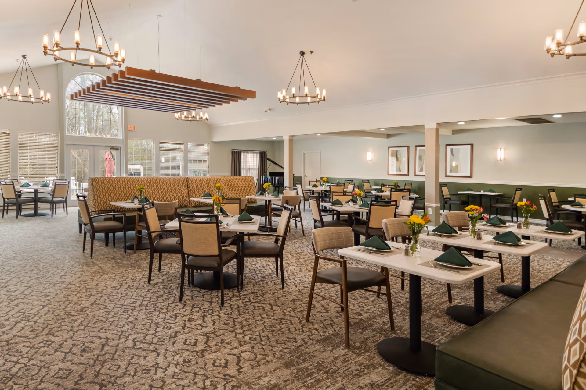 A spacious and well-lit dining room with multiple tables and chairs arranged neatly. Each table is set with green folded napkins and small vases of yellow flowers. The room features large windows allowing natural light, modern chandeliers hanging from the ceiling, and a patterned carpet floor. There is a mix of booth seating and individual chairs, with framed artwork on the walls.