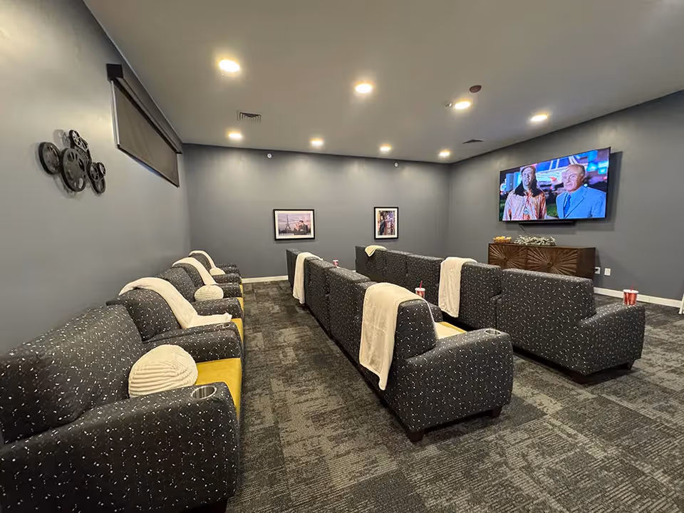 A cozy media room with rows of dark patterned armchairs, each with a light-colored throw blanket draped over the back. The room has gray walls, carpeted floor, and recessed ceiling lights. A flat-screen TV is mounted on the wall showing a scene with two people. There are framed pictures on the walls and a decorative wall piece resembling film reels.