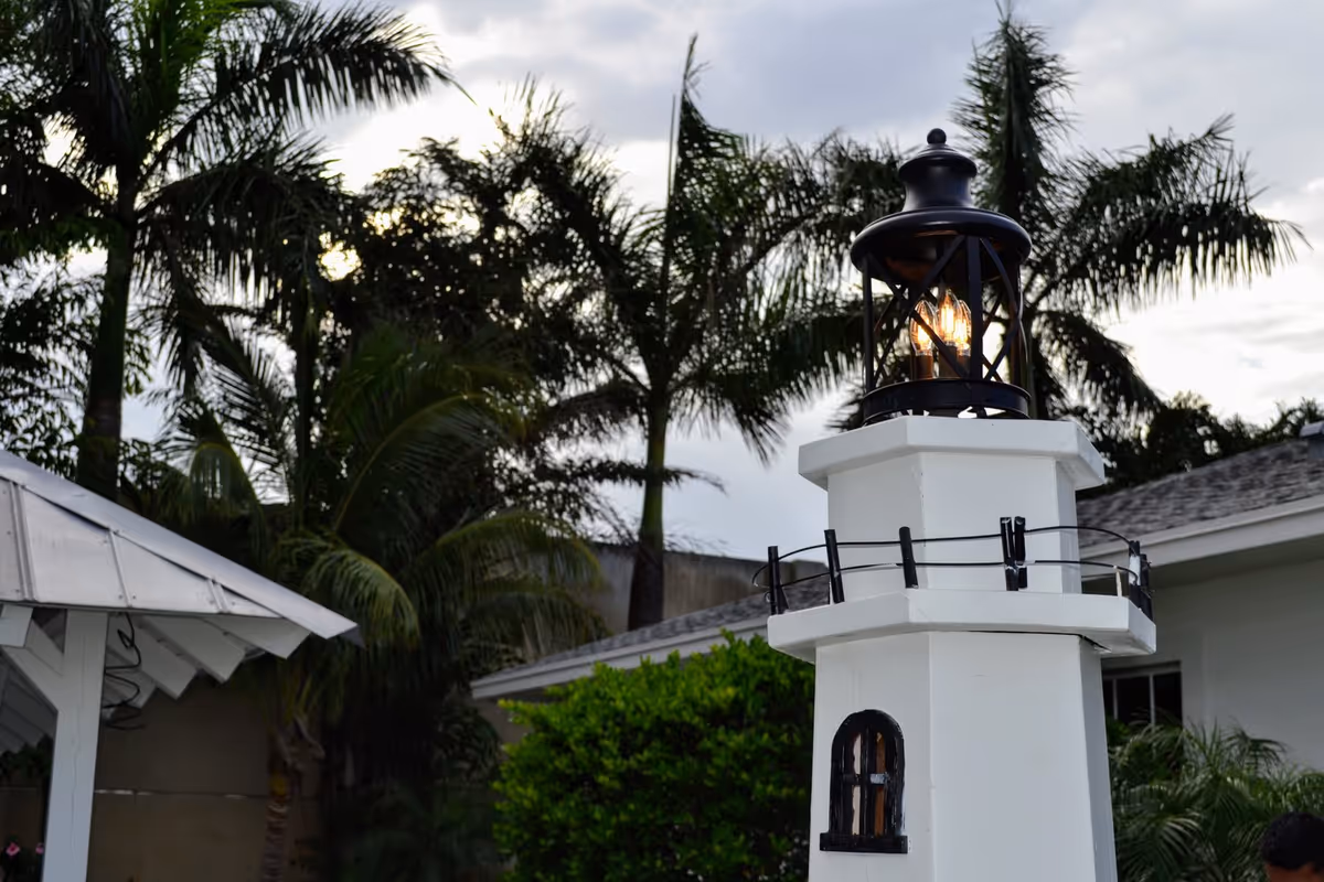 A white lighthouse-style outdoor lamp with a black lantern top and lit bulbs, surrounded by palm trees and greenery, with a building roof visible in the background under a cloudy sky.