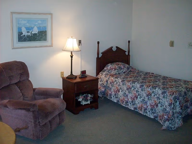 A small bedroom with a single bed covered in a floral bedspread, a wooden nightstand with a lamp and a small clock, a comfortable brown recliner chair, and a framed picture of two white chairs on a beach hanging on the wall.