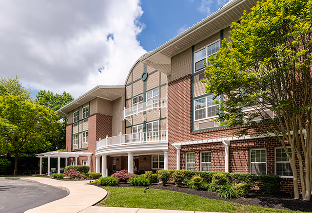 Exterior view of a three-story senior living facility building with brick and beige siding, surrounded by landscaped greenery and trees under a partly cloudy blue sky.