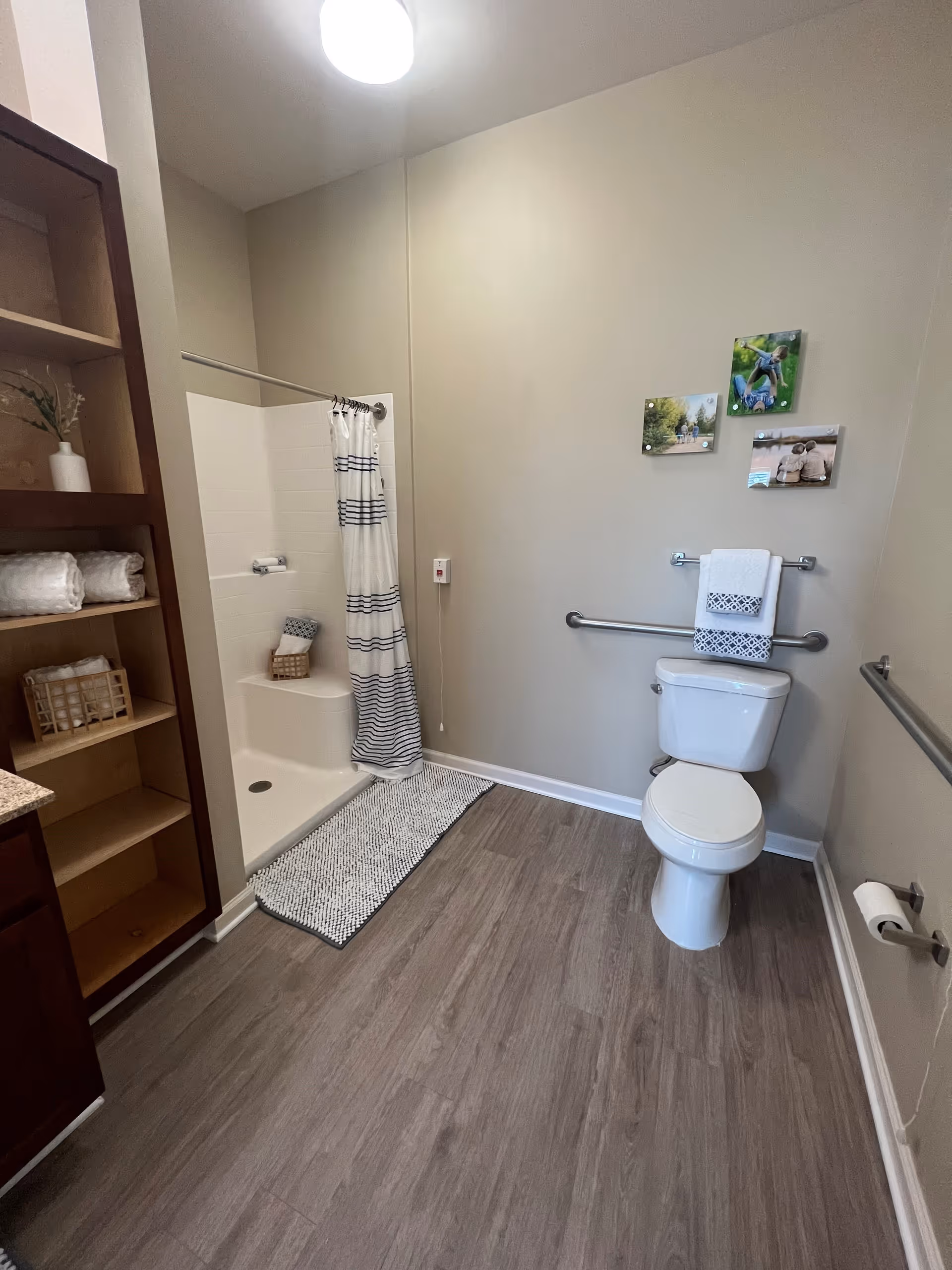 A clean and accessible bathroom with a white toilet, grab bars on the walls, a walk-in shower with a striped shower curtain, a gray bath mat, and wooden shelving holding towels and decorative items.