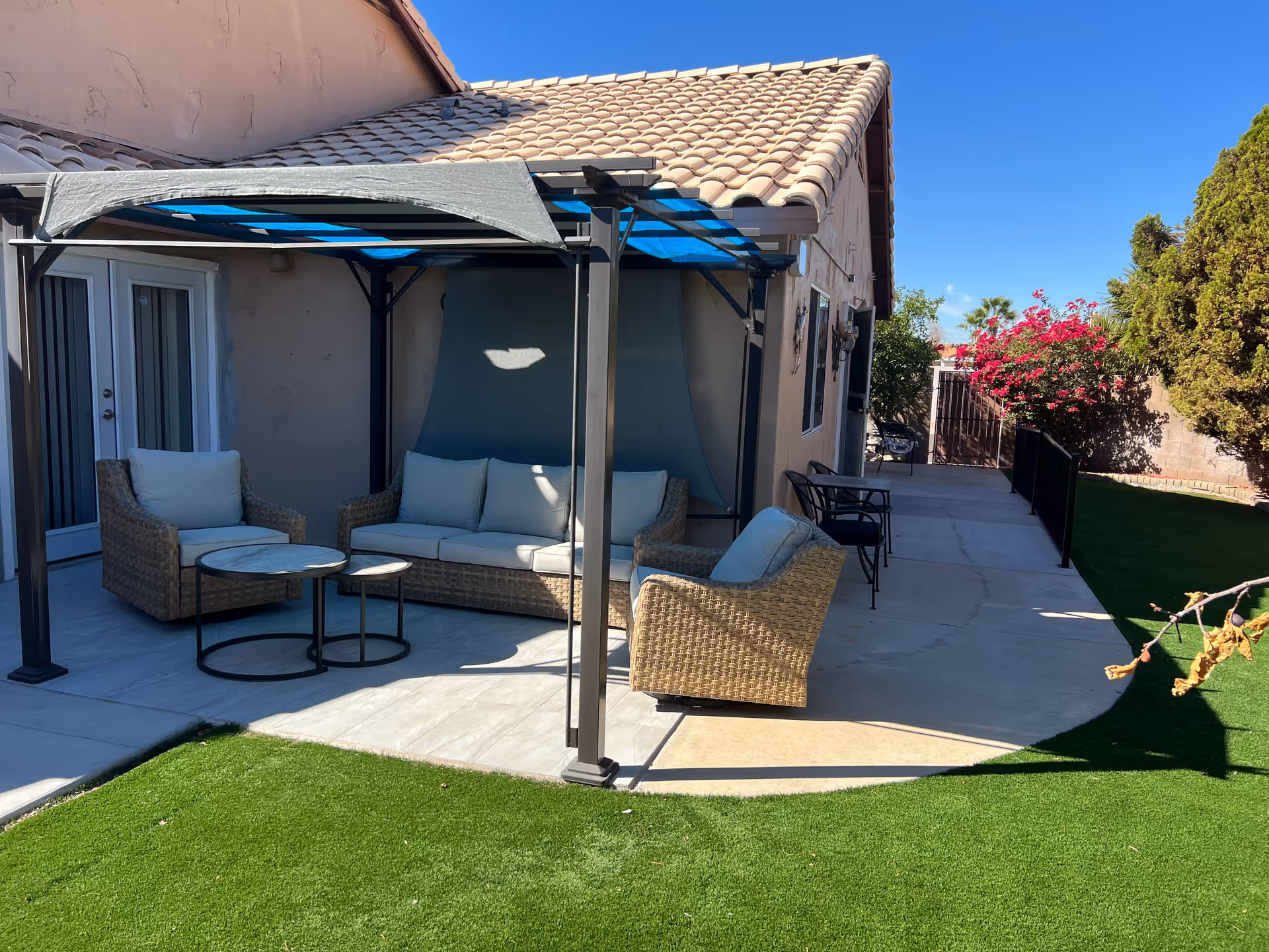 Outdoor patio area with wicker furniture including a sofa and two armchairs with cushions, a small round table, and a pergola providing partial shade. The patio is adjacent to a beige stucco building with a tiled roof. There is artificial green grass in the foreground and flowering bushes and trees in the background under a clear blue sky.
