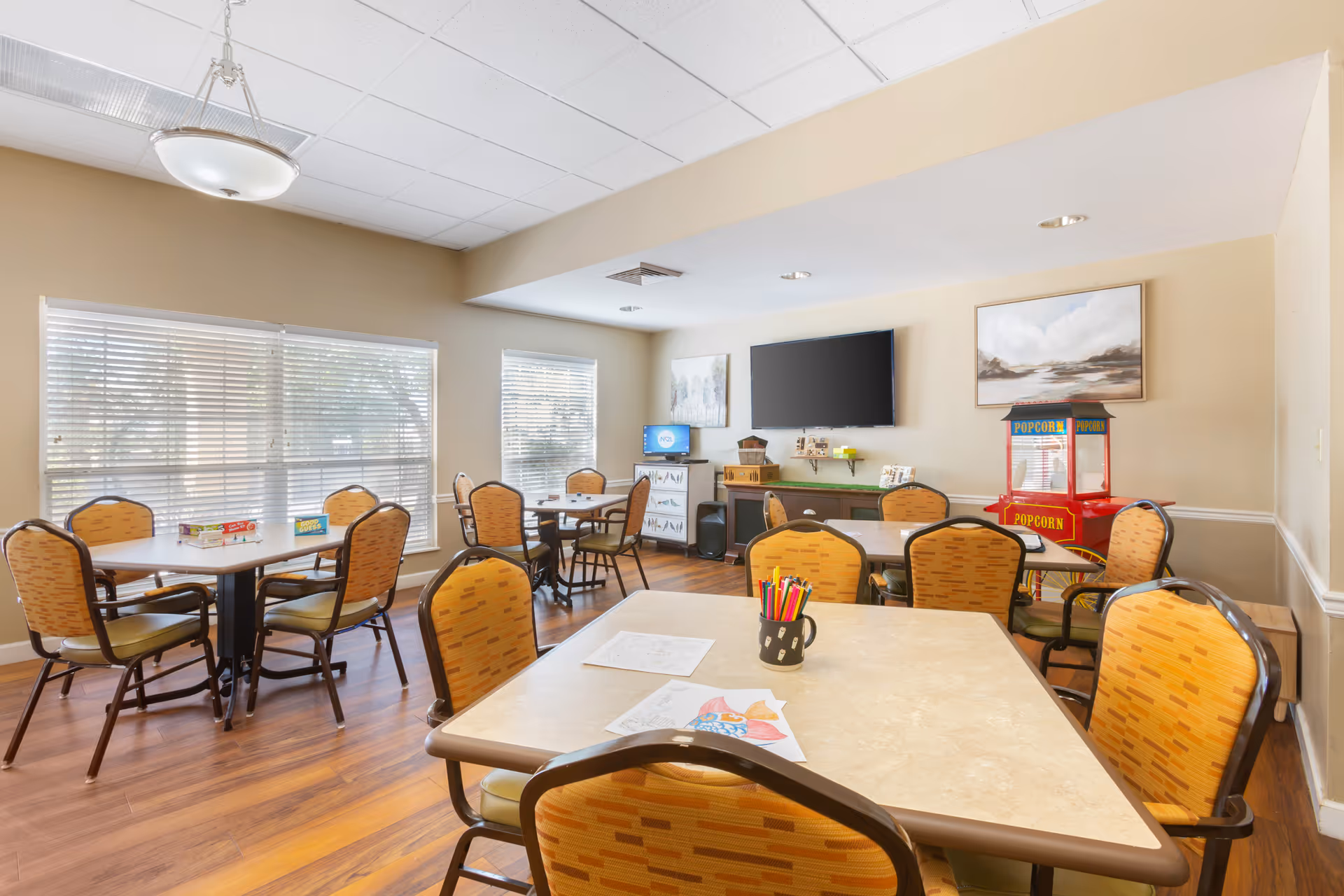 A bright activity room with several tables and orange patterned chairs. On the tables are coloring sheets, colored pencils, and board games. The room has large windows with blinds, a popcorn machine, a flat-screen TV mounted on the wall, and artwork hanging on the walls. The floor is wooden, and the ceiling has recessed lighting and a hanging light fixture.