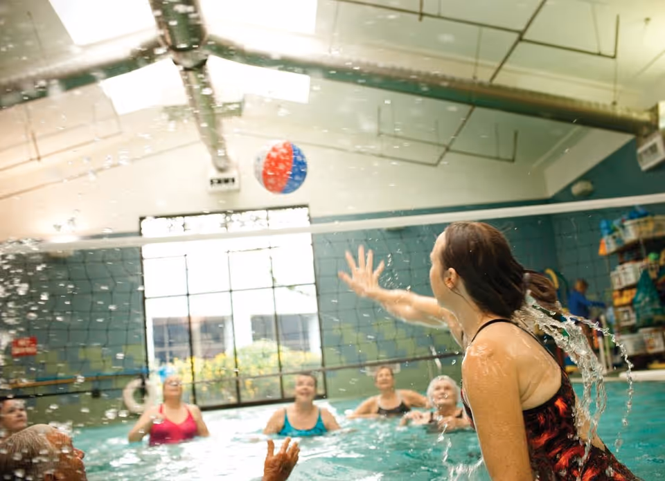 Seniors in an indoor swimming pool reaching and splashing as they play with a beach ball.