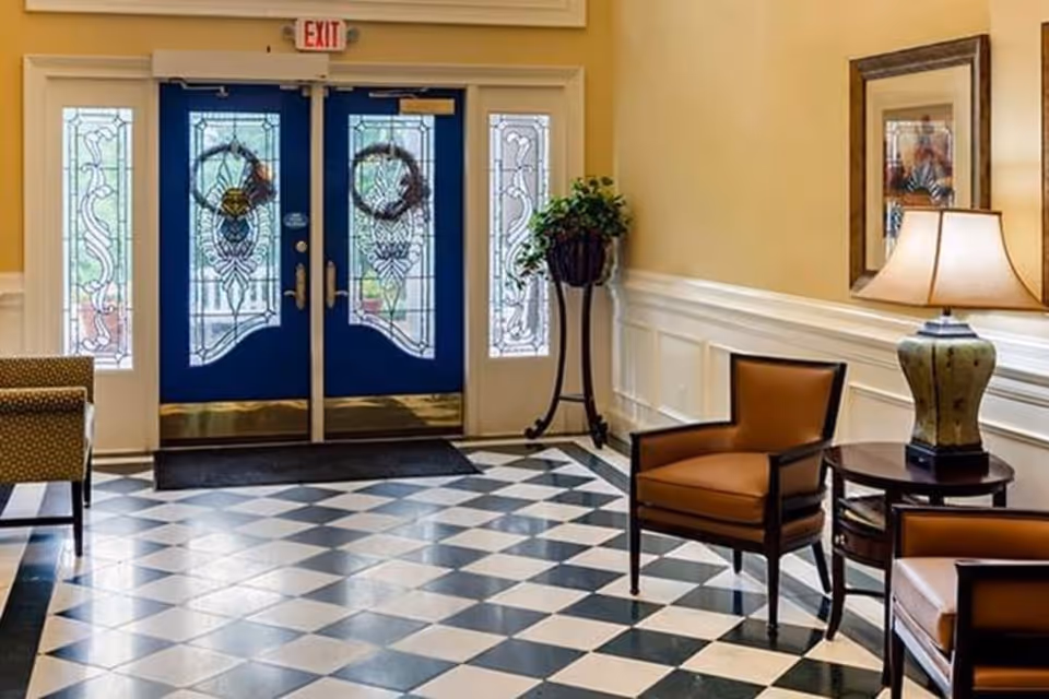 Entrance area of a facility with double blue doors featuring decorative glass panels and wreaths. The floor has a black and white checkered pattern. There are two brown armchairs and a round wooden table with a lamp on it. A potted plant stands near the door, and a framed picture hangs on the wall.