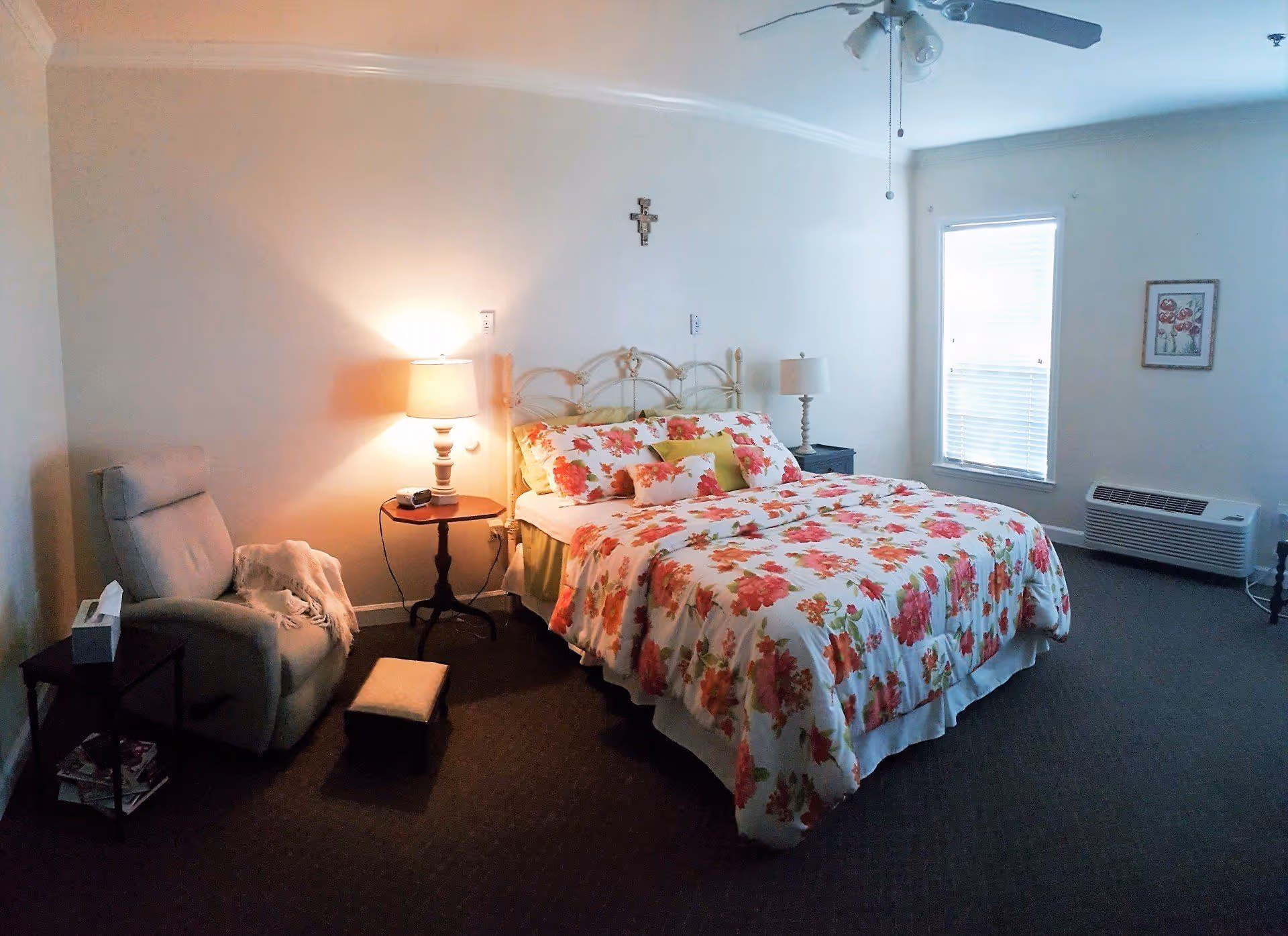 A cozy bedroom with a bed covered in floral bedding, two bedside tables with lamps, a comfortable armchair with a blanket, a small footstool, a window with blinds, a wall-mounted air conditioning unit, and a ceiling fan.