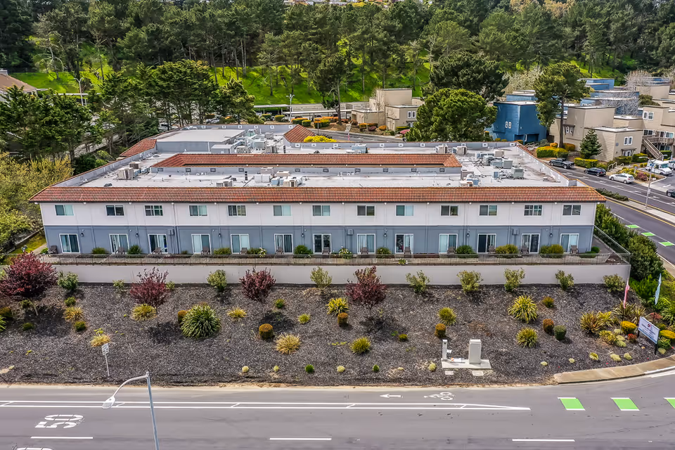 Aerial view of Serra Highlands Senior Living facility showing a two-story building with a tiled roof, multiple windows, and a landscaped area with shrubs and small trees in front. Surrounding the building are roads, parking areas, and additional residential buildings with greenery and trees in the background.