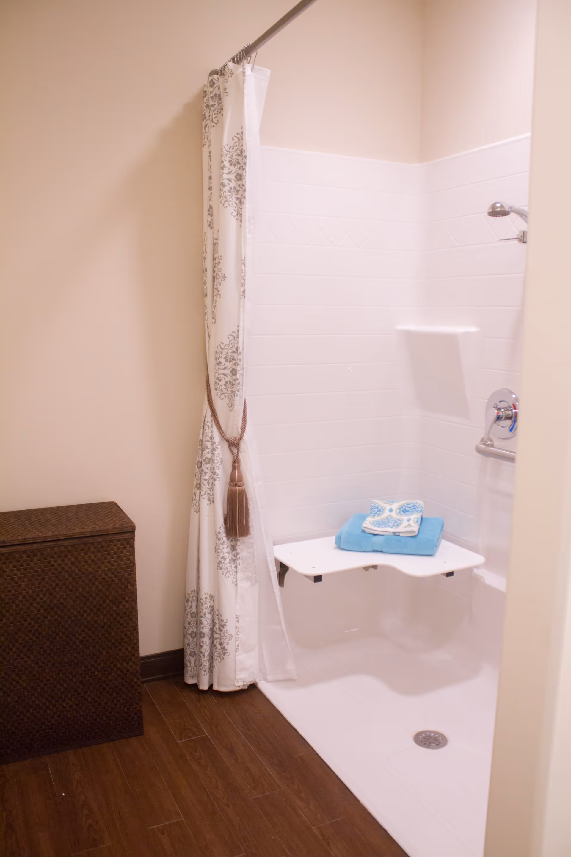A bathroom shower area with a white tiled wall and a fold-down shower seat holding a blue towel and a patterned washcloth. There is a decorative shower curtain tied back with a tassel, and a brown woven hamper is placed on the wooden floor outside the shower.