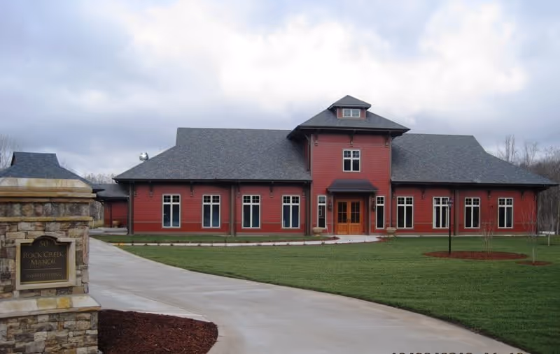 Front exterior of a red two-story building labeled Rock Creek Manor with a driveway, lawn, and a stone sign in the foreground.