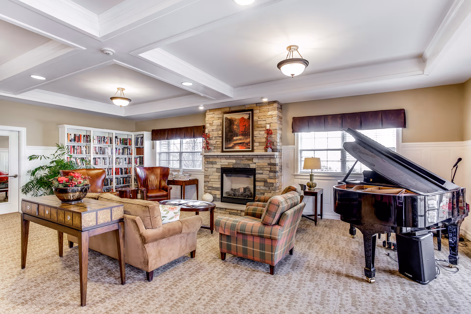 Bright communal living room with armchairs and sofas arranged around a stone fireplace, bookshelves, and a grand piano.