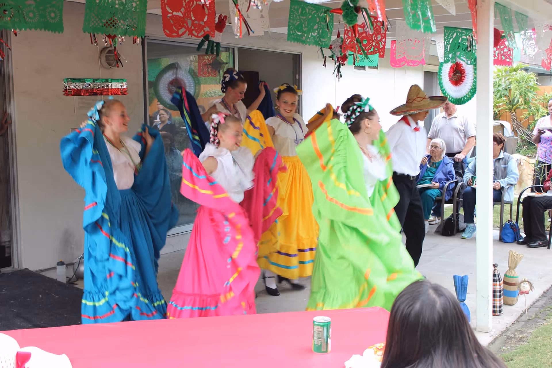 A group of young dancers dressed in colorful traditional Mexican attire perform a folkloric dance outdoors under a decorated patio. Several elderly people are seated and watching the performance. The patio is decorated with red, green, and white paper banners.