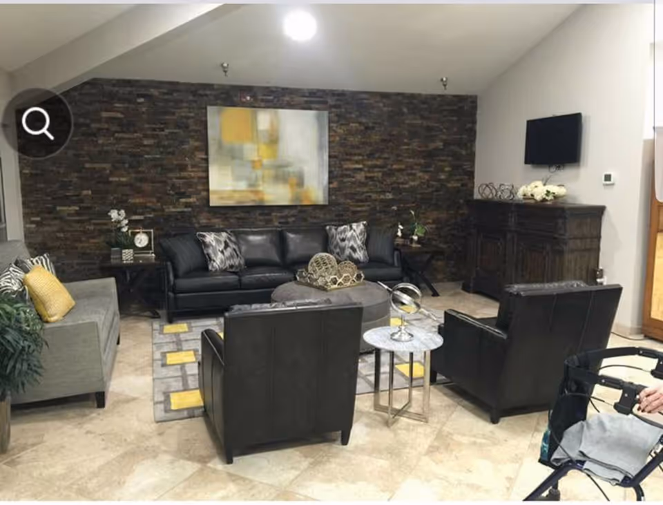 A cozy living room area with a black leather sofa, two black leather armchairs, and a gray fabric sofa with yellow and patterned cushions. The room features a stone accent wall with a modern abstract painting, a flat-screen TV mounted on the wall, a wooden cabinet, a round ottoman, and small side tables with decorative items. A walker is partially visible in the foreground.