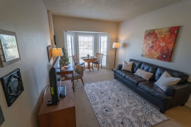 A cozy living room in a retirement community featuring a black leather sofa with decorative pillows, a patterned area rug, a wooden TV stand with a flat-screen television, a small round dining table with two chairs near a bay window, a floor lamp, and a large floral painting on the wall.
