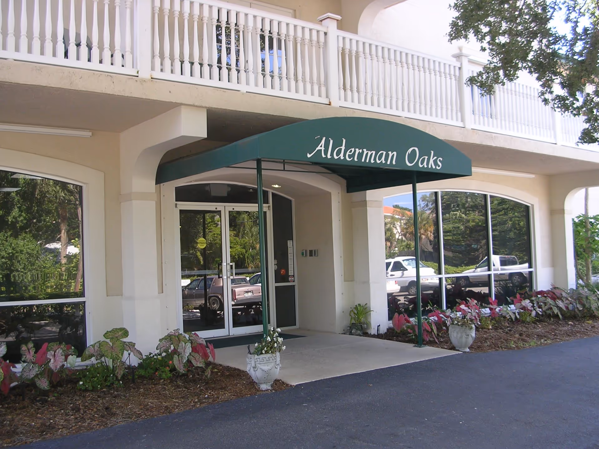 Entrance of Alderman Oaks Retirement Residence with a green awning displaying the facility name. The building has large windows reflecting parked cars and surrounding greenery. There are flower beds with plants on either side of the entrance.
