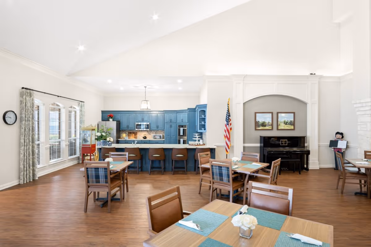 Spacious community dining area with tables and chairs, a blue kitchen island with barstools, a piano, and an American flag.